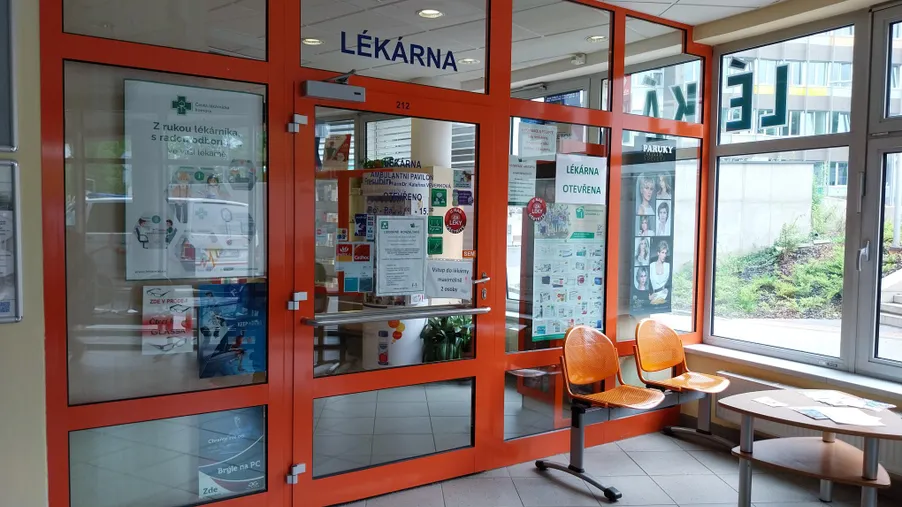 Vstup do moderní lékárny s oranžovým rámem a skleněnými dveřmi, poblíž jsou umístěny oranžové čekací židle.